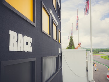 A large black scoreboard is prominently displayed with the word 'RACE' in bold white letters on its side. Surrounding the scoreboard are several yellow-highlighted rectangular slots. A woman in a white top and cap is standing on a balcony, looking towards a race track in the distance. The track is lined with green cones and several red and black flags are flying high. A vast cloudy sky and lush greenery form the backdrop.