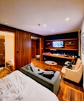 A cozy hotel room features a bed with white sheets in the foreground, adjacent to a gray sofa and tan armchair. The room has wood paneling and flooring, and a flat-screen TV mounted on the wall opposite a modern entertainment unit. The shelves hold various items including snacks and drinks, giving the room a lived-in feel. A bathroom is visible to the left, with part of a bathtub peeking through.