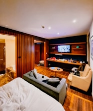 A cozy hotel room features a bed with white sheets in the foreground, adjacent to a gray sofa and tan armchair. The room has wood paneling and flooring, and a flat-screen TV mounted on the wall opposite a modern entertainment unit. The shelves hold various items including snacks and drinks, giving the room a lived-in feel. A bathroom is visible to the left, with part of a bathtub peeking through.