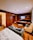 A cozy hotel room features a bed with white sheets in the foreground, adjacent to a gray sofa and tan armchair. The room has wood paneling and flooring, and a flat-screen TV mounted on the wall opposite a modern entertainment unit. The shelves hold various items including snacks and drinks, giving the room a lived-in feel. A bathroom is visible to the left, with part of a bathtub peeking through.