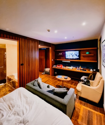 A cozy hotel room features a bed with white sheets in the foreground, adjacent to a gray sofa and tan armchair. The room has wood paneling and flooring, and a flat-screen TV mounted on the wall opposite a modern entertainment unit. The shelves hold various items including snacks and drinks, giving the room a lived-in feel. A bathroom is visible to the left, with part of a bathtub peeking through.