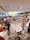 A bustling indoor event with people walking and interacting around various booths. The room has a modern design with bright lighting and geometric carpet patterns. Several informational materials and promotional stands are visible, indicating a possible career fair or exhibition.