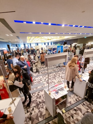 A bustling indoor event with people walking and interacting around various booths. The room has a modern design with bright lighting and geometric carpet patterns. Several informational materials and promotional stands are visible, indicating a possible career fair or exhibition.