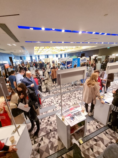 A bustling indoor event with people walking and interacting around various booths. The room has a modern design with bright lighting and geometric carpet patterns. Several informational materials and promotional stands are visible, indicating a possible career fair or exhibition.