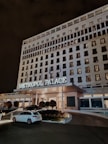 A large, multi-story hotel building with the name 'Metropol Palace' illuminated above the entrance. The facade is adorned with numerous rectangular windows, some of which are lit. At the entrance, there are a few trees and plants, as well as parked cars, including a white one in the foreground. The setting is at night, creating a serene and elegant atmosphere.