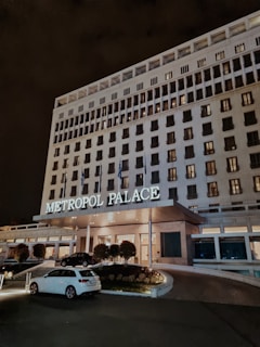 A large, multi-story hotel building with the name 'Metropol Palace' illuminated above the entrance. The facade is adorned with numerous rectangular windows, some of which are lit. At the entrance, there are a few trees and plants, as well as parked cars, including a white one in the foreground. The setting is at night, creating a serene and elegant atmosphere.