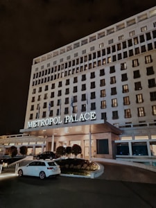 A large, multi-story hotel building with the name 'Metropol Palace' illuminated above the entrance. The facade is adorned with numerous rectangular windows, some of which are lit. At the entrance, there are a few trees and plants, as well as parked cars, including a white one in the foreground. The setting is at night, creating a serene and elegant atmosphere.
