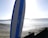 A paddleboard stands upright on a pebbly and sandy beach with calm waters stretching out to the horizon. The bright sun creates a reflective glare on the water, and a distant landmass is visible.