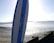 A paddleboard stands upright on a pebbly and sandy beach with calm waters stretching out to the horizon. The bright sun creates a reflective glare on the water, and a distant landmass is visible.