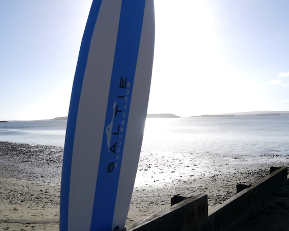 A paddleboard stands upright on a pebbly and sandy beach with calm waters stretching out to the horizon. The bright sun creates a reflective glare on the water, and a distant landmass is visible.