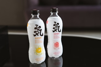Two bottles of sparkling water are placed on a reflective black surface. The bottles, labeled Genki, feature bold black and red text with images of citrus and peach fruits on them. Both bottles highlight '0 fat, 0 sugar, 0 calories' on the label.