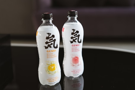 Two bottles of sparkling water are placed on a reflective black surface. The bottles, labeled Genki, feature bold black and red text with images of citrus and peach fruits on them. Both bottles highlight '0 fat, 0 sugar, 0 calories' on the label.