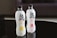 Two bottles of sparkling water are placed on a reflective black surface. The bottles, labeled Genki, feature bold black and red text with images of citrus and peach fruits on them. Both bottles highlight '0 fat, 0 sugar, 0 calories' on the label.