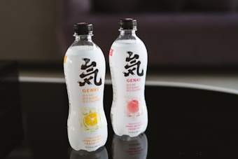 Two bottles of sparkling water are placed on a reflective black surface. The bottles, labeled Genki, feature bold black and red text with images of citrus and peach fruits on them. Both bottles highlight '0 fat, 0 sugar, 0 calories' on the label.