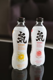 Two bottles of sparkling water are displayed, one with a citrus flavor and the other with a peach flavor. The bottles feature bold black lettering and symbols on a white label, with colorful fruit illustrations indicating their respective flavors. They are situated on a reflective black surface.