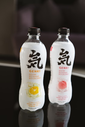 Two bottles of sparkling water are displayed, one with a citrus flavor and the other with a peach flavor. The bottles feature bold black lettering and symbols on a white label, with colorful fruit illustrations indicating their respective flavors. They are situated on a reflective black surface.