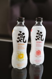 Two bottles of sparkling water are displayed, one with a citrus flavor and the other with a peach flavor. The bottles feature bold black lettering and symbols on a white label, with colorful fruit illustrations indicating their respective flavors. They are situated on a reflective black surface.