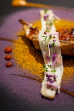 A gourmet dish artfully presented with a piece of grilled meat or poultry topped with a creamy white sauce garnished with microgreens and small purple flowers. The plate is artistically decorated with vibrant powders: a circle of purple powder and a semicircle of golden-yellow powder. Small dots of red sauce add contrast to the dish, and there is a dollop of an orange-colored sauce on the side.