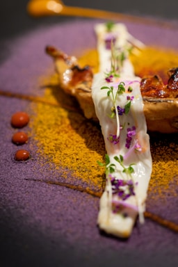 A gourmet dish artfully presented with a piece of grilled meat or poultry topped with a creamy white sauce garnished with microgreens and small purple flowers. The plate is artistically decorated with vibrant powders: a circle of purple powder and a semicircle of golden-yellow powder. Small dots of red sauce add contrast to the dish, and there is a dollop of an orange-colored sauce on the side.