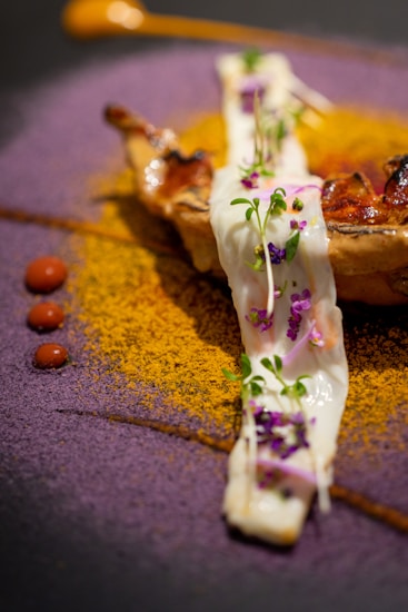 A gourmet dish artfully presented with a piece of grilled meat or poultry topped with a creamy white sauce garnished with microgreens and small purple flowers. The plate is artistically decorated with vibrant powders: a circle of purple powder and a semicircle of golden-yellow powder. Small dots of red sauce add contrast to the dish, and there is a dollop of an orange-colored sauce on the side.