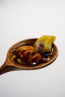 A wooden spoon holds a dark sauce with a glossy texture, along with a yellowish-green food item topped with a small purple flower.