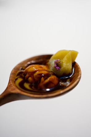 A wooden spoon holds a dark sauce with a glossy texture, along with a yellowish-green food item topped with a small purple flower.