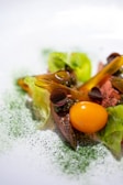 A gourmet dish featuring a vibrant yellow cherry tomato, surrounded by tender slices of cooked meat and lush green lettuce leaves. Garnished with dark berries and sprinkled finely with green herbs, adding a touch of elegance and freshness.