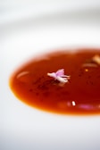 A vibrant red sauce garnished with a delicate pink edible flower is artfully presented on a white plate. The smooth surface of the sauce reflects light, adding a glossy texture to the presentation.