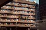 A multi-story brutalist-style building features rows of balconies adorned with plants and greenery. The concrete structure has a stark and utilitarian design, with red-brown accents on the window frames. The setting is urban, with neighboring buildings visible in the background and the scene illuminated by warm sunlight.