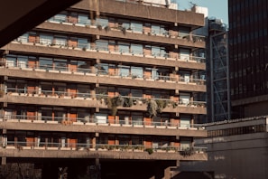 A multi-story brutalist-style building features rows of balconies adorned with plants and greenery. The concrete structure has a stark and utilitarian design, with red-brown accents on the window frames. The setting is urban, with neighboring buildings visible in the background and the scene illuminated by warm sunlight.