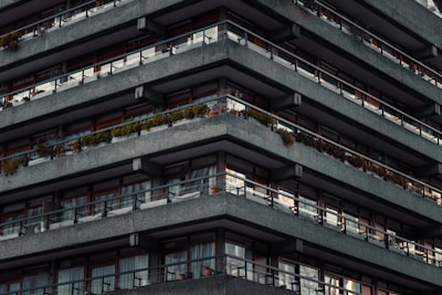 A multi-story building with repetitive, angular concrete balconies. Each level has railing lined with plants, and there are large windows or sliding doors behind the balconies. The facade appears uniform and modern, with a slightly industrial feel due to the concrete material.