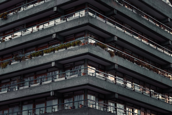 A multi-story building with repetitive, angular concrete balconies. Each level has railing lined with plants, and there are large windows or sliding doors behind the balconies. The facade appears uniform and modern, with a slightly industrial feel due to the concrete material.