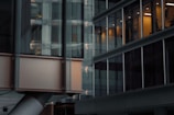A modern office building with large glass windows. The lighting inside creates a warm contrast against the exterior's cool tones. Light reflections can be seen on the windows, suggesting a multistory structure. The blurred column in the foreground suggests architectural elements.