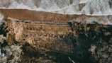 An aerial view of a coastline captures a meeting of land and sea, with waves crashing onto the shore creating a frothy boundary. The landscape is marked by grassy terrain and patches of trees, alongside an area showing signs of erosion. A row of vehicles is visible in the lower left corner, indicating human presence.