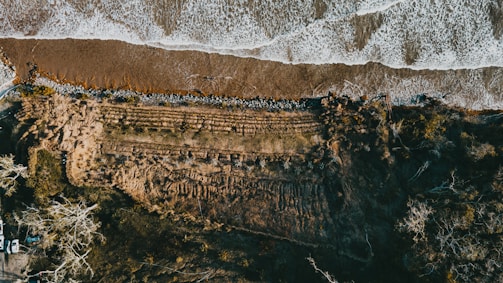 An aerial view of a coastline captures a meeting of land and sea, with waves crashing onto the shore creating a frothy boundary. The landscape is marked by grassy terrain and patches of trees, alongside an area showing signs of erosion. A row of vehicles is visible in the lower left corner, indicating human presence.