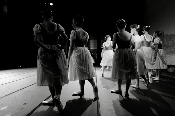A group of dancers in traditional ballet costumes stand on stage, surrounded by a dark theater setting. The costumes are reminiscent of classical ballet attire with light, flowing skirts and fitted bodices. The scene conveys a sense of anticipation and grace as the dancers wait in formation.