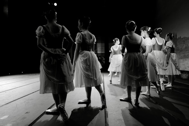 A group of dancers in traditional ballet costumes stand on stage, surrounded by a dark theater setting. The costumes are reminiscent of classical ballet attire with light, flowing skirts and fitted bodices. The scene conveys a sense of anticipation and grace as the dancers wait in formation.