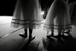 Dancers in delicate, layered skirts standing on a dimly lit stage, casting soft shadows on the floor.
