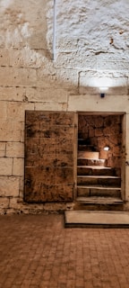 A dimly lit stone room with a wooden door slightly open, revealing a short staircase leading upwards. The walls and floor are made of stone, with some areas illuminated by small lights. The texture of the stone is rough and aged.
