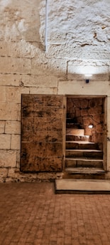 A dimly lit stone room with a wooden door slightly open, revealing a short staircase leading upwards. The walls and floor are made of stone, with some areas illuminated by small lights. The texture of the stone is rough and aged.