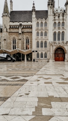 A historic building with Gothic architectural features, including pointed arches and ornate details. The facade is made of light-colored stone, and the structure is adjacent to an expansive tiled plaza with geometric patterns. A modern black vehicle and a canopy are visible by the side of the building.