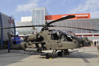 A military helicopter is on display outdoors in front of a modern building with a glass facade. The helicopter appears complex, with notable features such as rotors and equipment attached to its body. There are several people in military attire nearby, observing the helicopter.