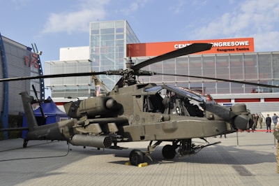 A military helicopter is on display outdoors in front of a modern building with a glass facade. The helicopter appears complex, with notable features such as rotors and equipment attached to its body. There are several people in military attire nearby, observing the helicopter.