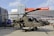A military helicopter is on display outdoors in front of a modern building with a glass facade. The helicopter appears complex, with notable features such as rotors and equipment attached to its body. There are several people in military attire nearby, observing the helicopter.