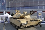 A military tank is displayed outdoors in front of a modern glass building. The tank is tan-colored with visible tracks and a large cannon. Surrounding the tank are informational displays and white tents.