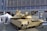 A military tank is displayed outdoors in front of a modern glass building. The tank is tan-colored with visible tracks and a large cannon. Surrounding the tank are informational displays and white tents.