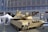 A military tank is displayed outdoors in front of a modern glass building. The tank is tan-colored with visible tracks and a large cannon. Surrounding the tank are informational displays and white tents.