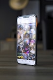 A smartphone displaying an Instagram profile with several thumbnail images on its screen, showing various lifestyle and technology-related pictures. The phone is placed on a tabletop in a dimly lit room with blurred background elements.