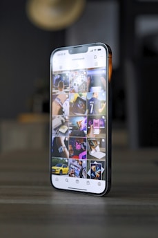 A smartphone displaying an Instagram profile with several thumbnail images on its screen, showing various lifestyle and technology-related pictures. The phone is placed on a tabletop in a dimly lit room with blurred background elements.
