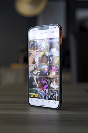 A smartphone displaying an Instagram profile with several thumbnail images on its screen, showing various lifestyle and technology-related pictures. The phone is placed on a tabletop in a dimly lit room with blurred background elements.